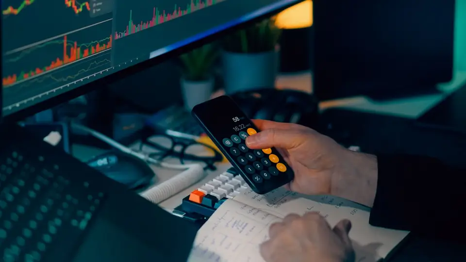 Pessoa usando a calculadora do celular diante de gráficos de mercado na tela do computador